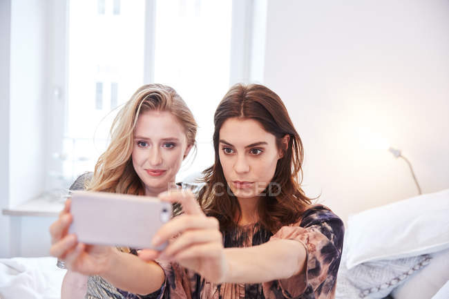 Две молодые женщины сидят на кровати и делают селфи на смартфоне. — стоковое фото