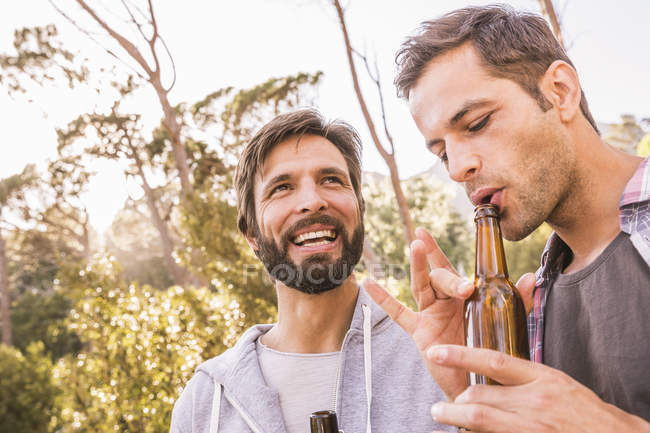 Two men blowing into beer bottle whilst camping in forest, Deer Park, Cape Town, South Africa — Stock Photo