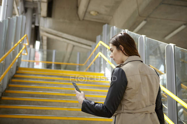 Visão traseira da jovem empresária lendo textos de smartphones na escada, Londres, Reino Unido — Fotografia de Stock