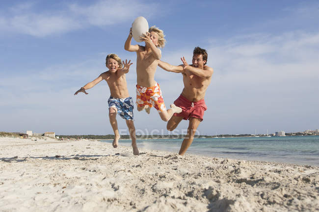 Menino saltando com bola de rúgbi perseguido por irmão e pai na praia, Maiorca, Espanha — Fotografia de Stock