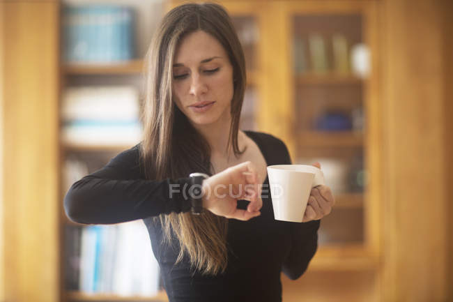 Junge Frau zu Hause, hält Heißgetränk in der Hand, schaut auf die Uhr — Stockfoto