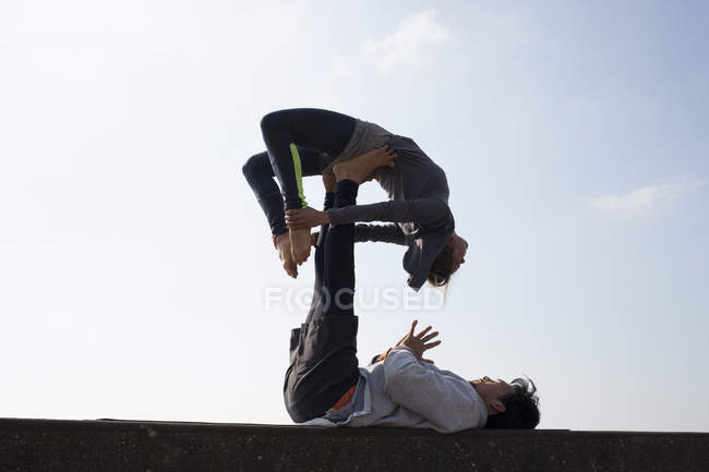 Siluetas hombre y mujer practicando yoga acrobático en la pared contra el cielo azul - foto de stock