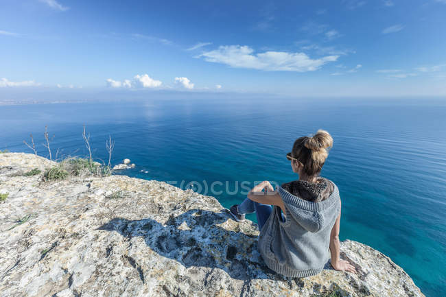 Veduta posteriore di una giovane donna seduta sulla scogliera che guarda verso l'oceano, Cagliari, Sardegna, Italia — Foto stock
