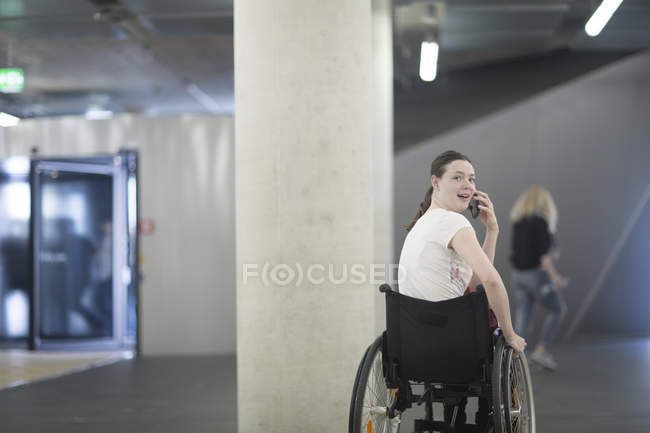 Mujer joven usando silla de ruedas en el estacionamiento subterráneo hablando en el teléfono inteligente - foto de stock