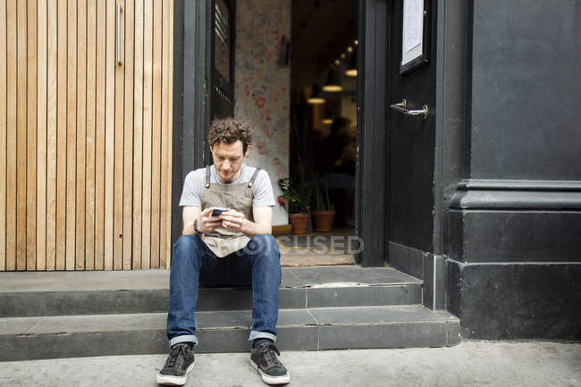 Waiter taking a break on cafe step looking at smartphone — Stock Photo