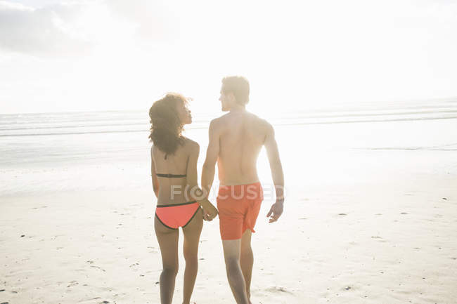 Vista traseira do jovem casal em roupa de banho passeando na praia iluminada pelo sol, Cidade do Cabo, Cabo Ocidental, África do Sul — Fotografia de Stock