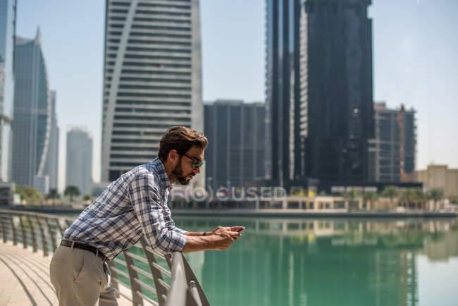 Young man leaning against waterfront railings reading smartphone texts, Dubai, United Arab Emirates — Stock Photo