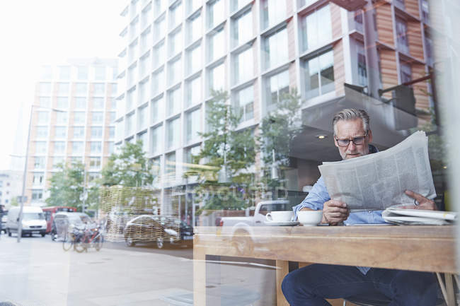 Reifer Mann sitzt im Café, liest Zeitung, Straße spiegelt sich im Fenster — Stockfoto