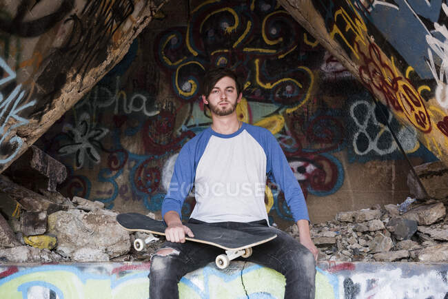 Portrait of young male skateboarder sitting on graffiti wall at ruined mine — Stock Photo