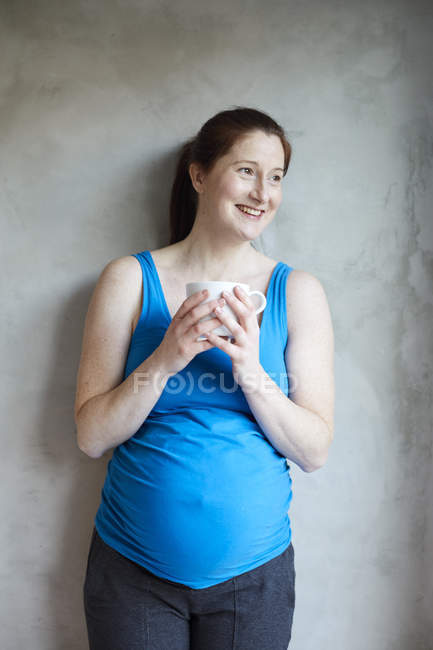 Mulher grávida encostada à parede segurando xícara de café olhando para longe sorrindo — Fotografia de Stock