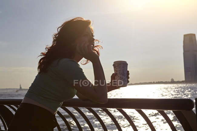 Joven turista apoyada en barandilla frente al mar con café, Manhattan, Nueva York, EE.UU. - foto de stock
