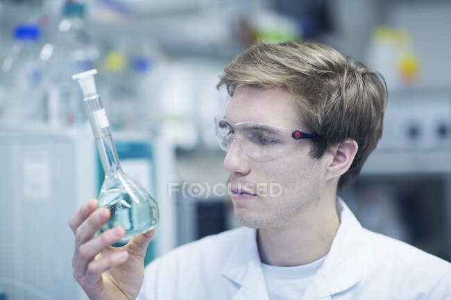 Male scientist holding up and looking at flask in lab — Stock Photo