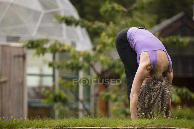 Mujer madura practicando yoga hacia adelante pliegue en eco lodge jardín - foto de stock