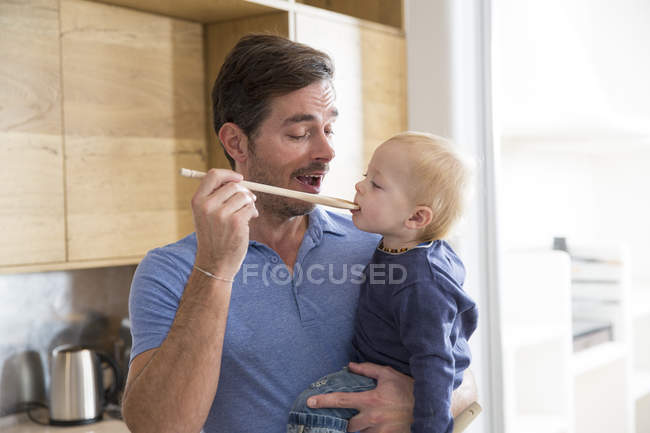 Чоловік годує маленького сина дерев'яною ложкою на кухні — стокове фото