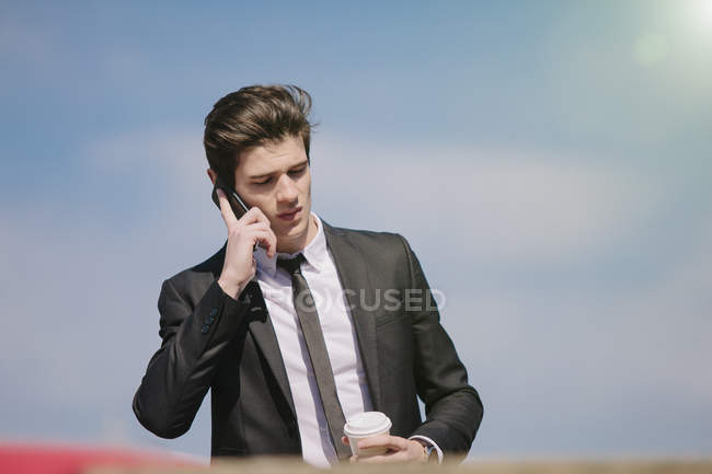 Молодой бизнесмен разговаривает по смартфону и пьет кофе на вынос — стоковое фото