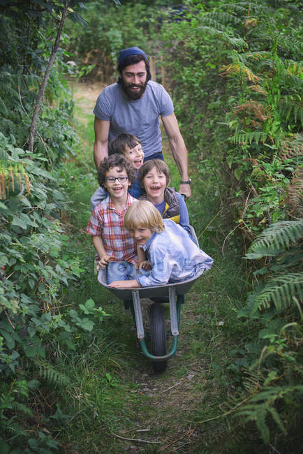 Mittlerer erwachsener Mann schiebt Schubkarre mit lächelnden Jungen in Garten — Stockfoto
