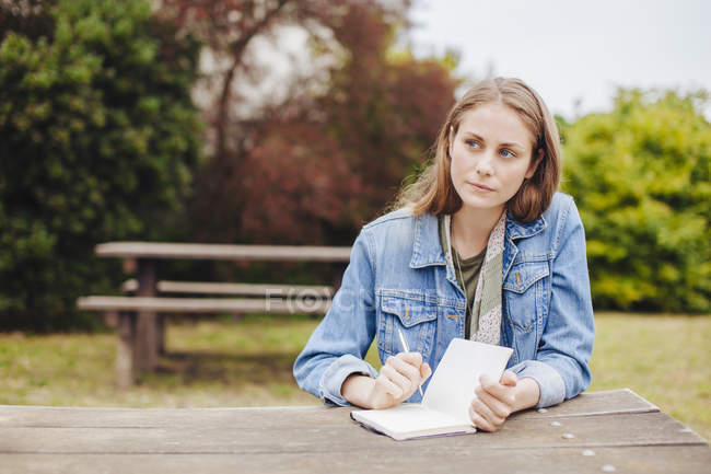 Junge Frau sitzt an Picknickbank im Park und schreibt in Notizbuch — Stockfoto