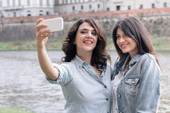 Pareja lesbiana usando smartphone para tomar selfie junto al río Arno, Florencia, Toscana, Italia - foto de stock