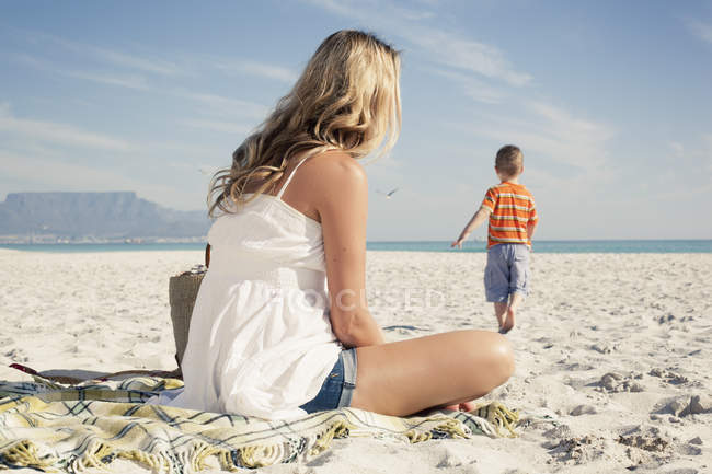 Madre viendo mientras joven hijo jugando en la playa - foto de stock