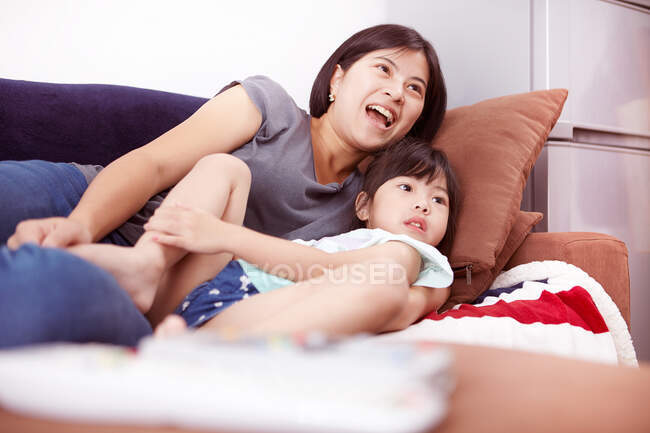 Junge chinesische Mutter und Tochter liegen auf dem Sofa und schauen zu Hause gemeinsam fern — Stockfoto