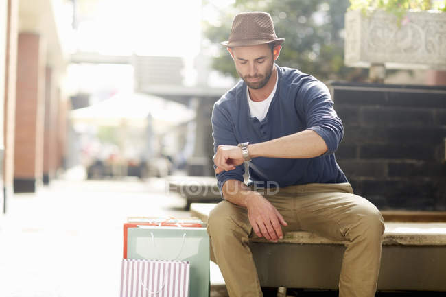 Hombre adulto sentado en el asiento, mirando el reloj, bolsas de compras a su lado - foto de stock