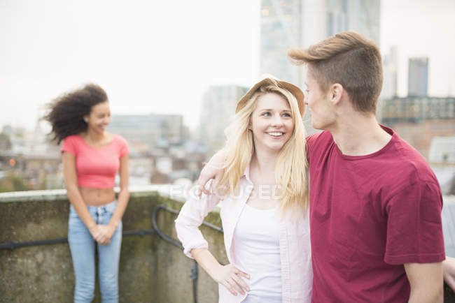 Junges Paar mit umschlungenen Armen, junge Frau im Hintergrund — Stockfoto