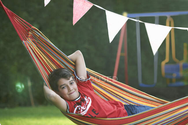Портрет уверенного мальчика, лежащего в полосатом садовом гамаке с руками за головой — стоковое фото