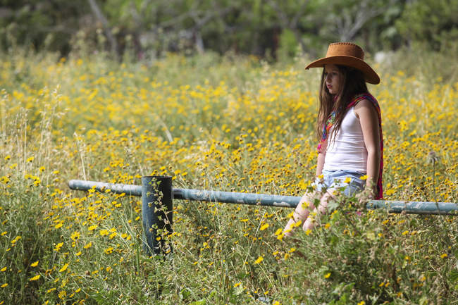Sullen girl in cowboy hat sitting on fence in field — Stock Photo
