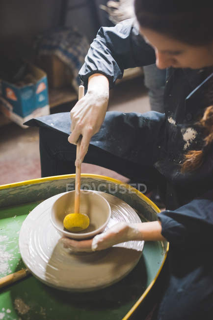 Young woman sitting at pottery wheel forming clay pot with diddler — Stock Photo