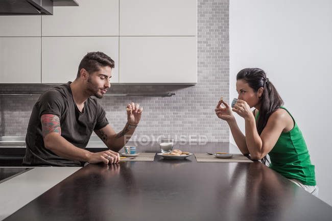 Casal jovem em frente uns dos outros tomando café da manhã na cozinha — Fotografia de Stock