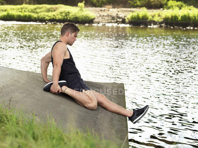 Young man exercising beside river, stretching leg — Stock Photo