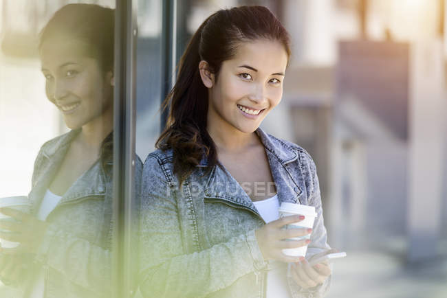 Ritratto di giovane donna con caffè da asporto e smartphone appoggiata all'edificio del parco — Foto stock