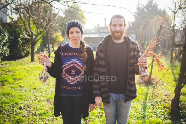 Paar steht mit frischen Knoblauchzwiebeln im Garten und blickt lächelnd in die Kamera — Stockfoto