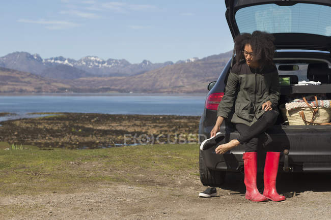 Mujer adulta sentada en una bota de auto poniéndose zapatos, Loch Eishort, Isla de Skye, Hébridas, Escocia - foto de stock