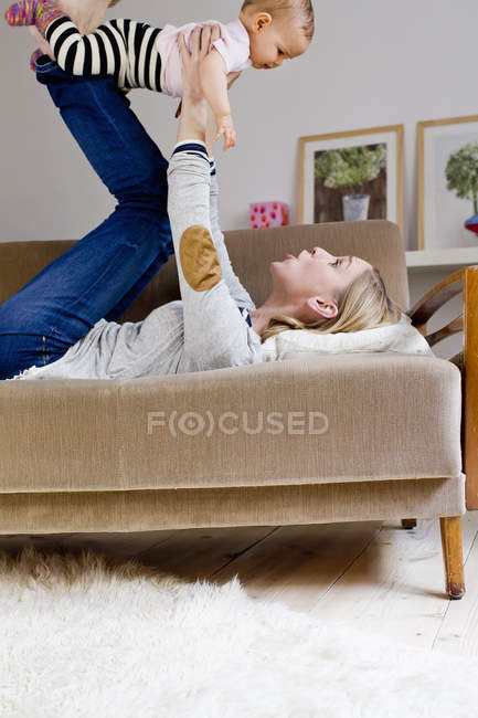 Mère couchée sur le canapé, tenant bébé fille dans l'air — Photo de stock