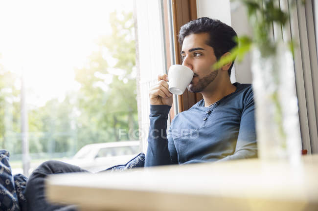 Young man sitting in front of window drinking coffee, looking away — Stock Photo