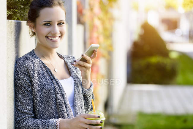 Retrato de una mujer joven apoyada en la pared del parque usando un smartphone - foto de stock