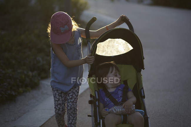 Девушка толкает младшую сестру в коляске через парк на закате — стоковое фото