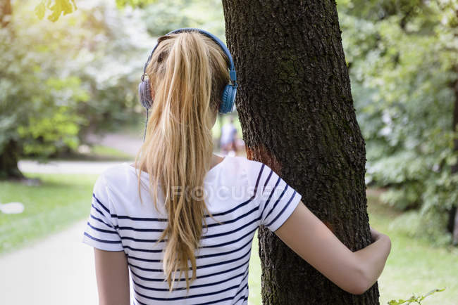 Rückansicht einer jungen Frau mit Pferdeschwanz, Kopfhörer tragend, Arm um Baum, wegschauend — Stockfoto