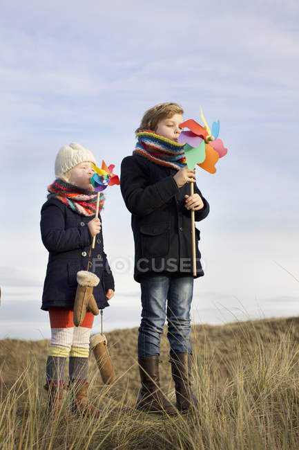 Irmão e irmã soprando em moinhos de vento de papel na costa — Fotografia de Stock