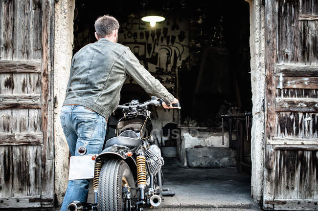 Задний вид человека, толкающего мотоцикл в сарай — стоковое фото