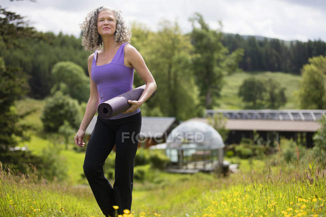 Зрелая женщина с ковриком для йоги ходит в эко-домике поле — стоковое фото