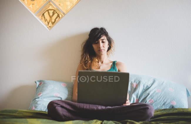Front view of mid adult woman sitting cross legged on bed using laptop computer — Stock Photo