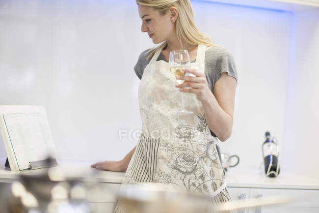 Молода жінка п'є келих білого вина під час читання книги рецептів на кухні — стокове фото