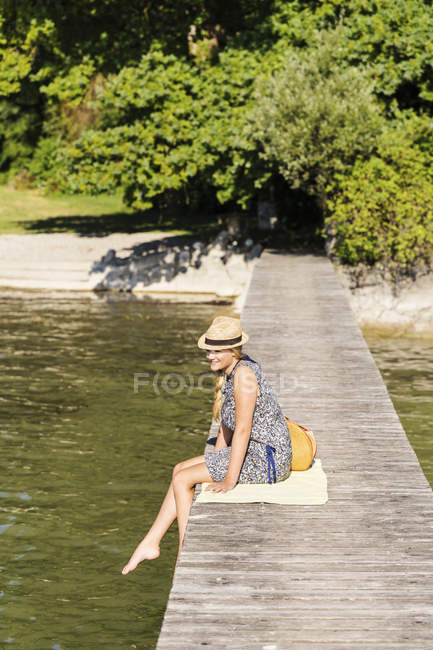 Side view of young woman sitting on wood pier wearing panama hat looking away, Schondorf, Ammersee, Bavaria, Germany — Stock Photo