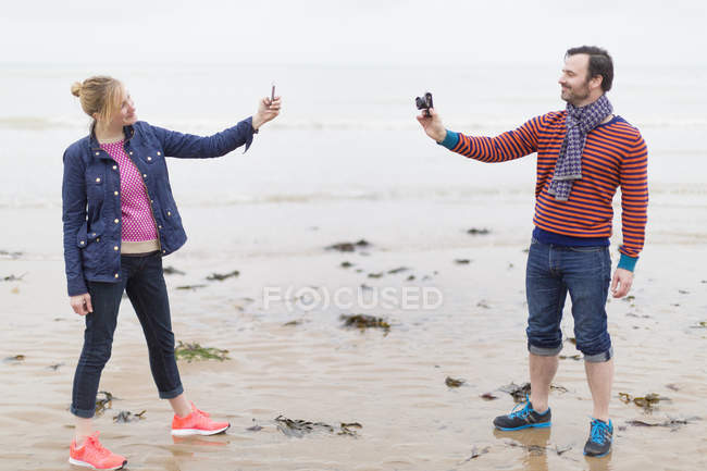 Пара на мокром пляже фотографируют друг друга с камерой и смартфоном — стоковое фото