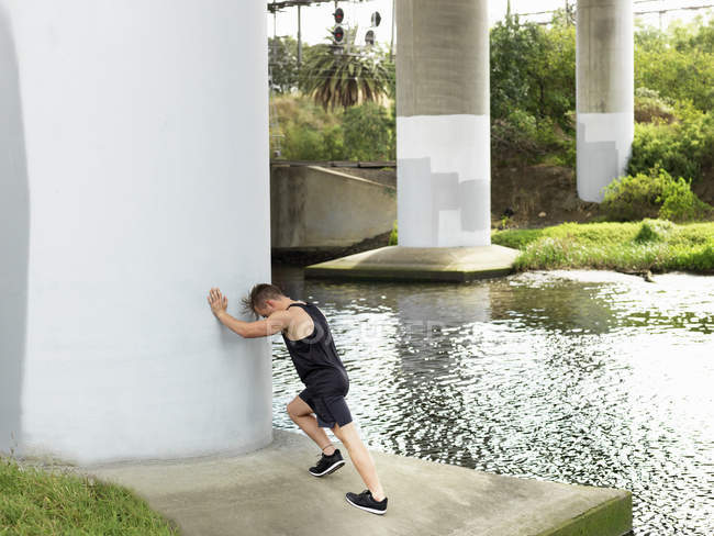 Young man exercising beside river, leaning against column, stretching — Stock Photo