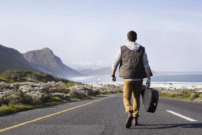 Visão traseira do jovem com caixa de guitarra andando na estrada costeira — Fotografia de Stock