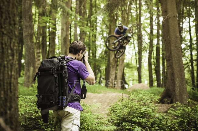 Joven fotógrafo masculino fotografiando ciclista de montaña en el bosque - foto de stock