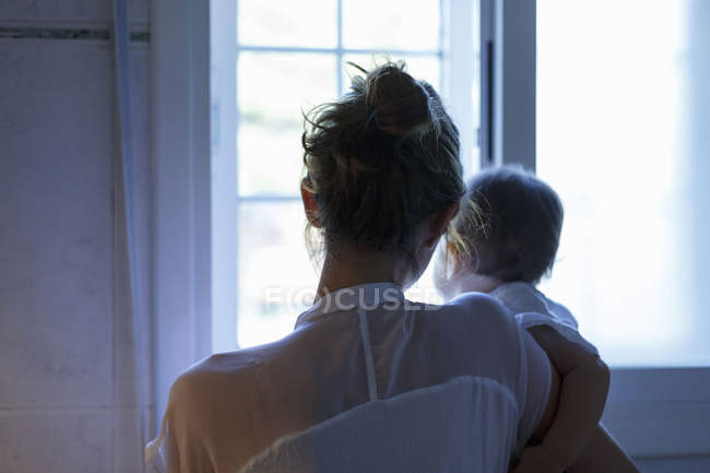 Vista trasera de la mujer adulta y la hija bebé mirando por la ventana de la sala de estar - foto de stock